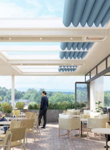 A modern rooftop patio with tables and chairs, people relaxing, reading, and enjoying beverages, overlooking lush greenery under a clear sky.