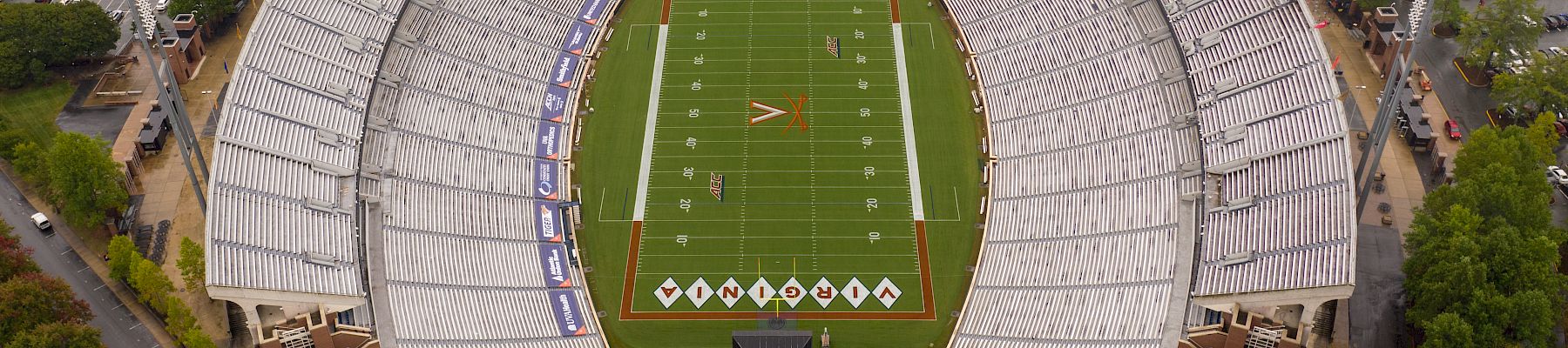 An aerial view of a large football stadium with a green field, white yard lines, and surrounding seating filled with light-colored seats, all amid trees and roads.